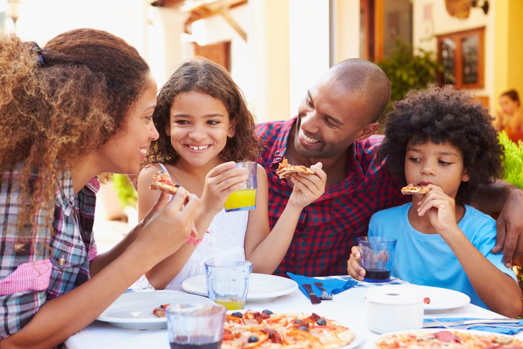 Family Eating Meal At Outdoor Restaurant Together, Smiling, woman eating a piece of pizza, girl eating a slice of pizza and drinking orange juice, man smiling and boy eating pizza