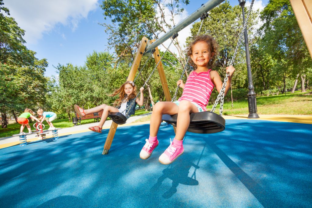 Two beautiful little girls swinging on swings on playground with smile on sunny summer day