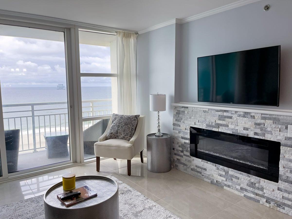 picture showing a wide fireplace with stone around it, a balcony in the background with the ocean visible and a chair that looks inviting at Seaside Resort in a 2-bedroom